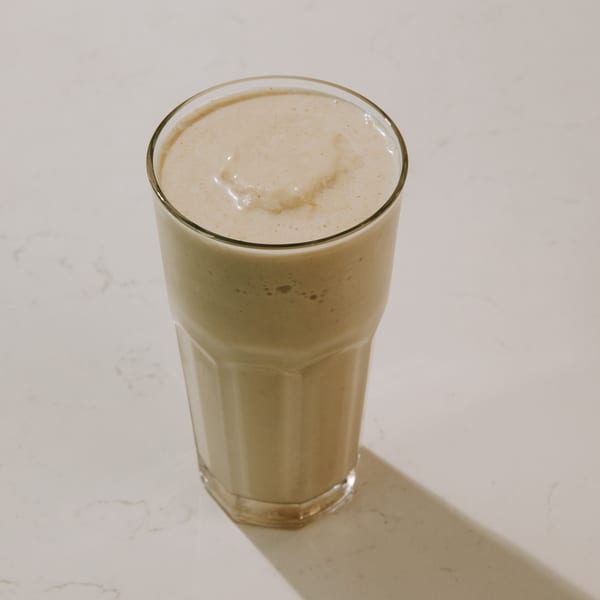 Tall glass filled with a light brown smoothie on a white marble surface.
