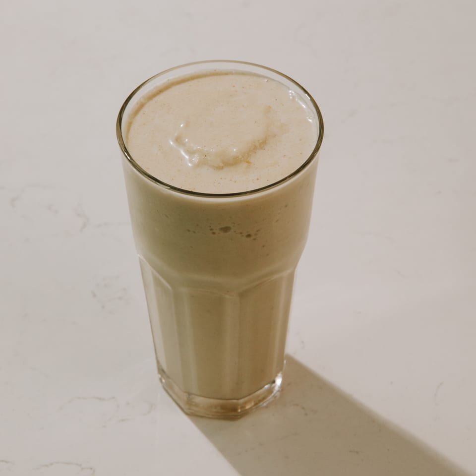 Tall glass filled with a light brown smoothie on a white marble surface.