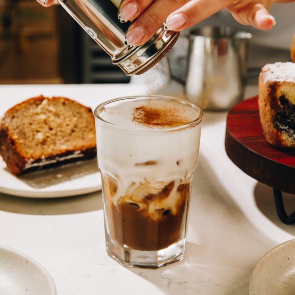 A hand sprinkles cinnamon on a glass of iced coffee with cream, next to slices of cake.