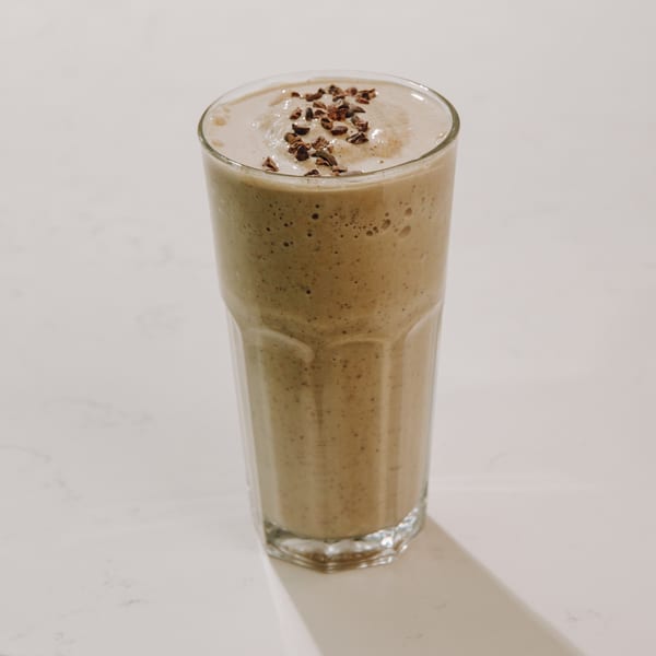 A tall glass of light brown smoothie, topped with dark chocolate nibs, on a white surface.