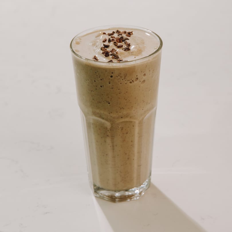 A tall glass of light brown smoothie, topped with dark chocolate nibs, on a white surface.