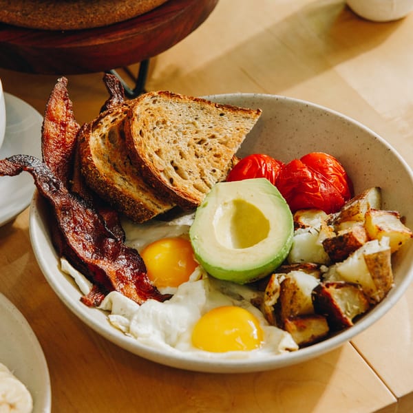 A breakfast bowl with bacon, fried eggs, avocado, roasted tomatoes, potatoes, and toast.