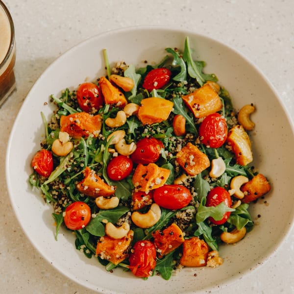 A vibrant salad with roasted tomatoes, butternut squash, cashews, quinoa, and arugula in a white bowl.