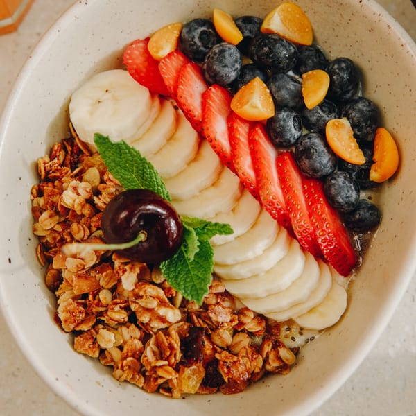 A bowl of granola with sliced bananas, strawberries, blueberries, ground cherries, and a mint sprig.