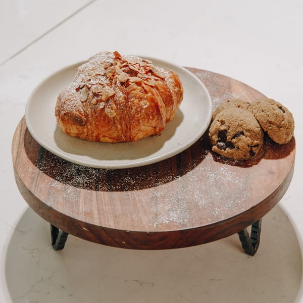 Almond croissant with powdered sugar on a white plate, with three chocolate chip cookies on a wooden stand.