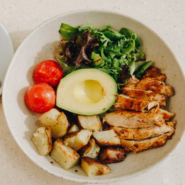 A healthy meal with sliced chicken, roasted potatoes, avocado, cherry tomatoes, and mixed greens.